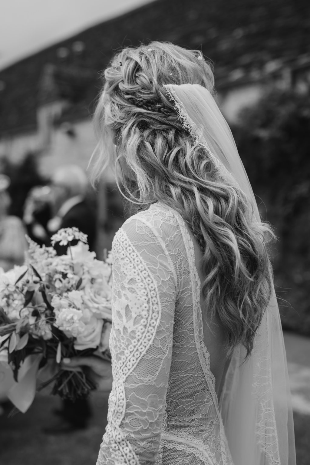 A black-and-white photo of a bride from behind, showing intricate lace on her dress and a styled bridal braid with veil and flowers.